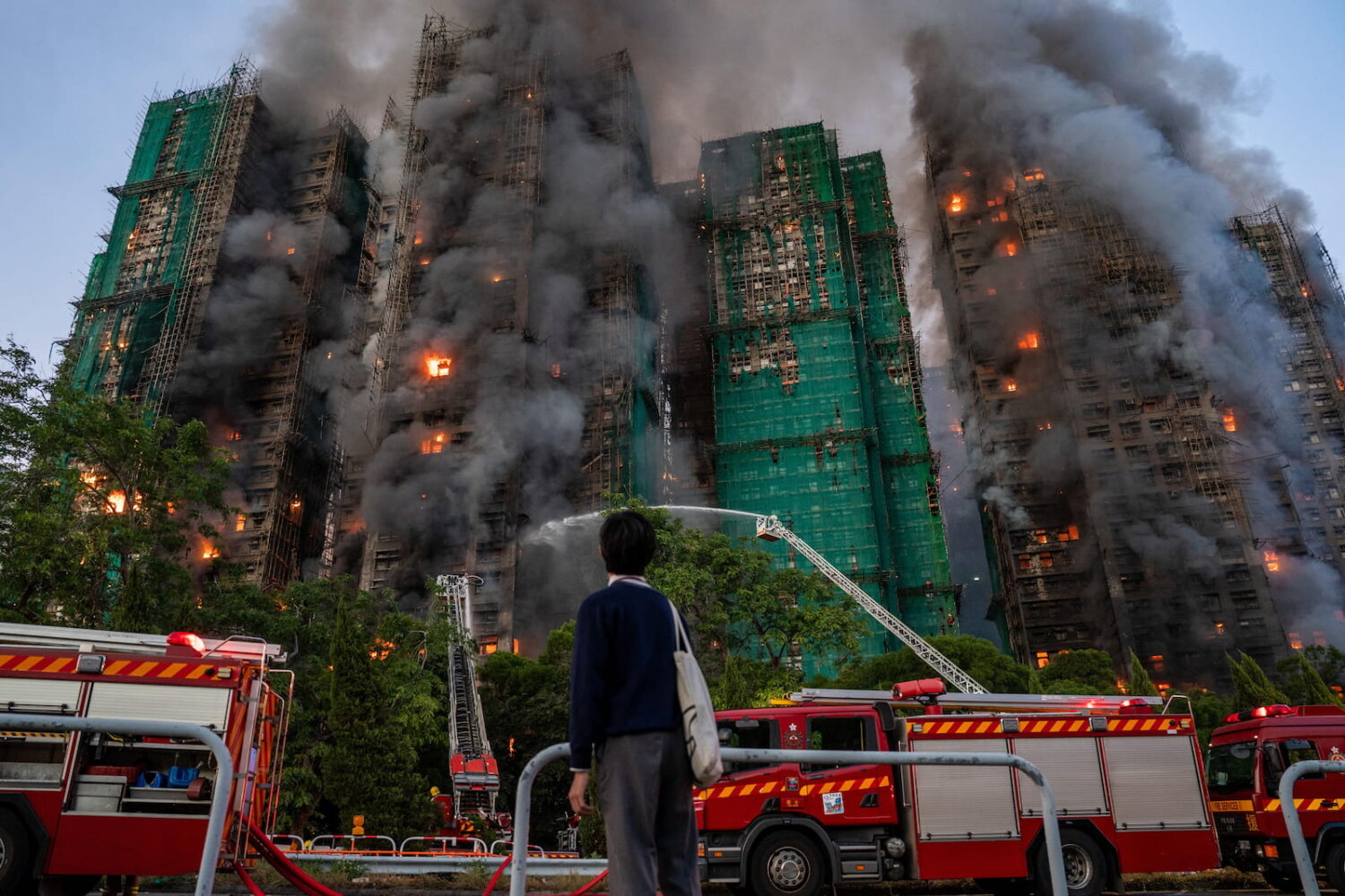 Una persona observa de pie el incendio en Hong Kong mientras varias torres cubiertas con andamios de bambú arden y desprenden gruesas columnas de humo, con camiones de bomberos y mangueras intentando controlar las llamas en la base de los edificios